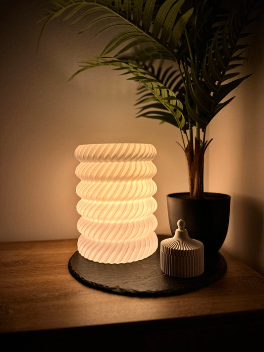 Moderne 3D-gedruckte weiße Tischlampe mit geschwungenem Wellenmuster und warmem Ambientelicht, neben einem Pflanzentopf auf Holzuntergrund.