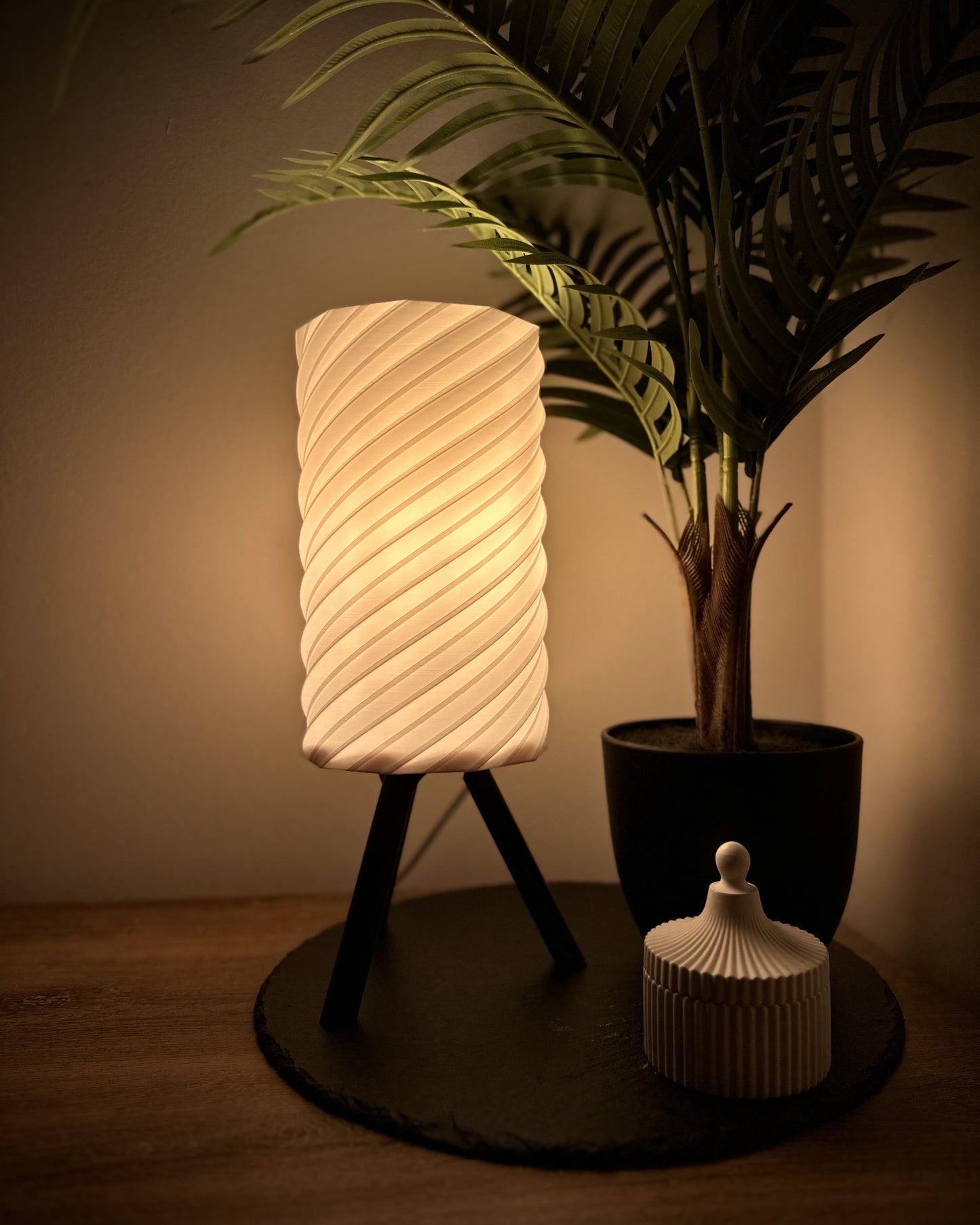 Dimmbare Tischlampe mit warmweißem Licht, spiralförmigem Lampenschirm und Dreibein-Gestell, dekoriert mit Pflanze und Dose.