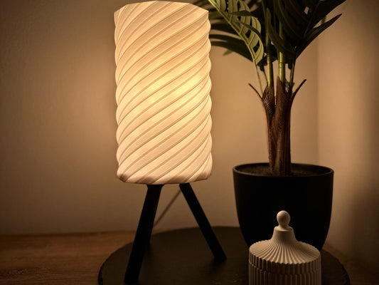 Dimmbare Tischlampe mit warmweißem Licht, spiralförmigem Lampenschirm und Dreibein-Gestell, dekoriert mit Pflanze und Dose.