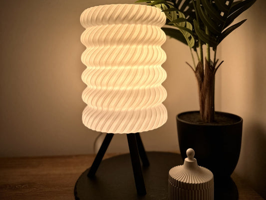 Stilvolle dimmbare Tischlampe mit warmweißem Licht, Spiralschirm und Dreibein, umgeben von Pflanzen und dekorativer Dose