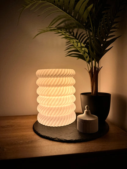 Moderne 3D-gedruckte weiße Tischlampe mit geschwungenem Wellenmuster und warmem Ambientelicht, neben einem Pflanzentopf auf Holzuntergrund.