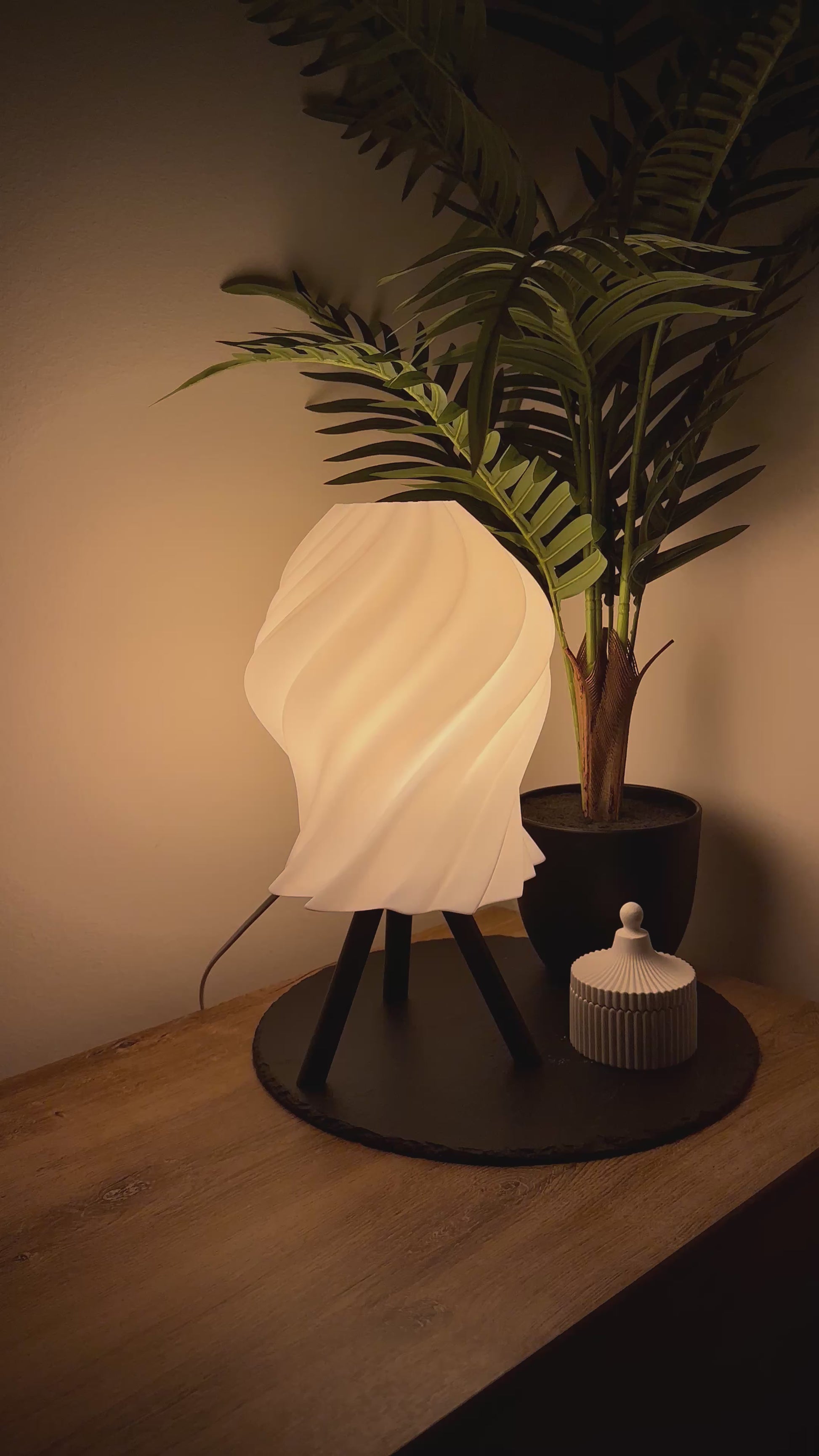 Moderne, dimmbare Lampe mit geschwungenem Schirm und Dreibeinfuß, begleitet von einer stilvollen Dose