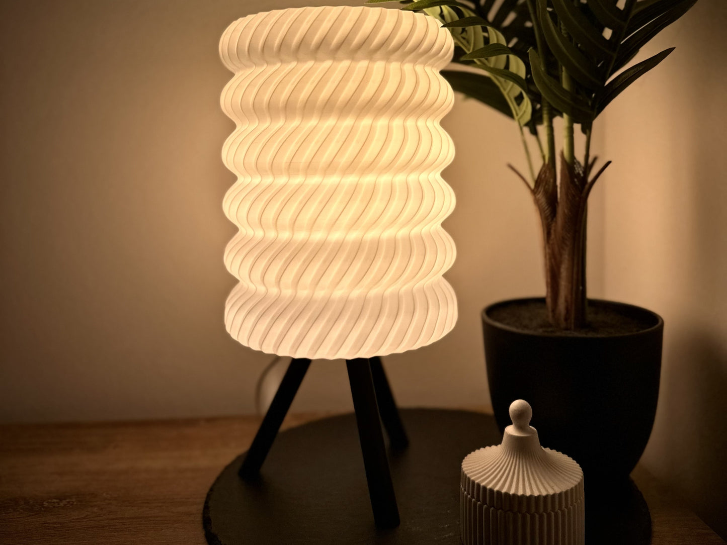 Stilvolle dimmbare Tischlampe mit warmweißem Licht, Spiralschirm und Dreibein, umgeben von Pflanzen und dekorativer Dose