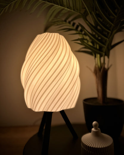 Tischlampe mit spiralig geschwungenem Schirm, dimmbar, warmweiß, Dreibeinfuß, ergänzt durch Pflanze und kleine Dose