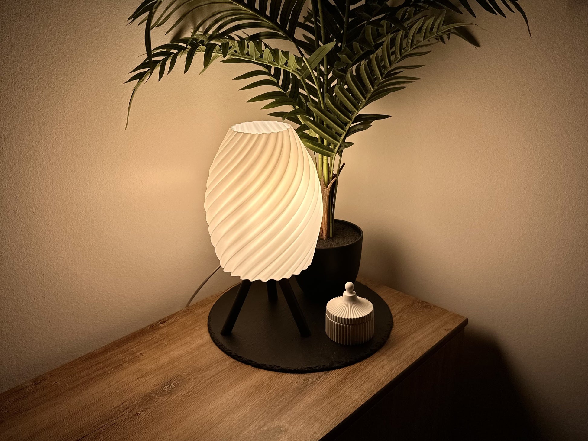 Moderne, dimmbare Lampe mit weißem Spiraldesign, stehend auf drei Beinen, neben einer Zimmerpflanze und einer Dose