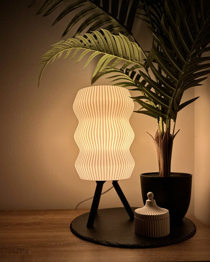 Moderne Dimmbare Tischlampe mit wellenförmigem Schirm auf dreibeinigem Ständer, warmweißes licht, neben Pflanze und Deko